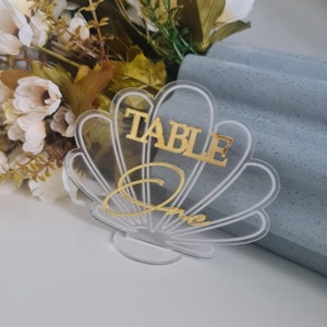 Op de afbeelding: Duidelijke acryl tafelnummer met een schelpontwerp. Het nummer "een" is geschreven in gouden schrift en de woorden "Tafel Eén" zijn in gouden blokletters.