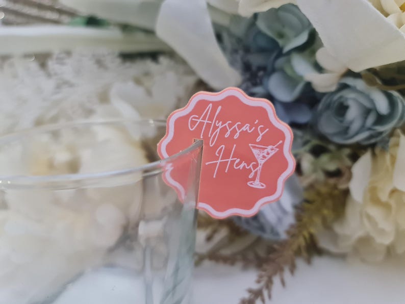 May include: A coral drinks charm with a scalloped edge, featuring the text "Alyssa's Hens" and a martini glass illustration. The charm is attached to a clear glass, with a floral arrangement in the background.