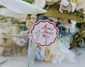 May include: A white and red scalloped-edge tag with the words "Gems Hens" and a cocktail illustration. The tag is attached to a clear glass and surrounded by a floral arrangement with white, cream, and blue flowers.