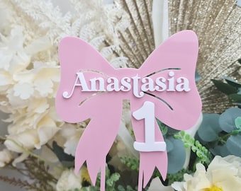 May include: A pink acrylic cake topper in the shape of a bow with the name "Anastasia" in white script. A white number "1" is below the name. The topper is designed for a first birthday celebration.