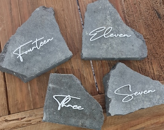 May include: Four gray stone table number holders with white cursive text. The numbers are Fourteen, Eleven, Three, and Seven. The stones are on a wooden surface.