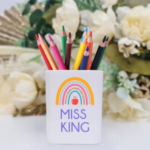 May include: White ceramic pencil holder with a rainbow design and the text "Miss King". The rainbow has a red apple in the centre and the words "Believe, Dream, Inspire, Motivate, Teach, Love, Learn, Encourage".