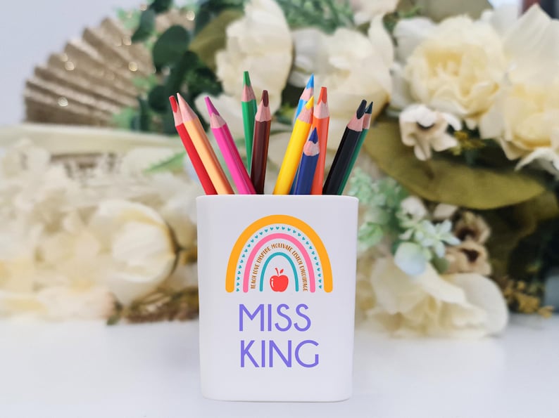 May include: White ceramic pencil holder with a rainbow design and the text "Miss King". The rainbow has a red apple in the center and the words "Believe, Dream, Inspire, Motivate, Teach, Love, Learn, Encourage".