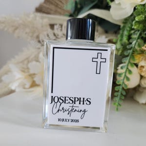 Può includere: Una bottiglia di vetro trasparente con tappo nero. L'etichetta recita "JOSEPH'S Christening" con un simbolo a croce e la data "10 LUGLIO 2025". La bottiglia è rettangolare e poggia su una superficie bianca.