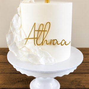 May include: A white cake with gold lettering spelling "Athraa" is presented on a white cake stand. The cake features a white, ruffled decoration on its side. A gold candle is placed on top of the cake.