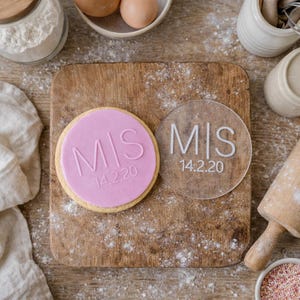 May include: A round cookie with pink frosting and the letters "MIS" and the date "14.2.20" embossed on top. Next to it is a clear acrylic circle with the same text. The items are on a wooden cutting board.