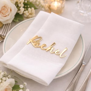Puede incluir: Una placa dorada con el nombre "Rachel" en escritura cursiva descansa sobre una servilleta de lino blanco doblada. La servilleta está colocada sobre un plato blanco con un borde verde fino, parte de una mesa. Flores y una vela desenfocadas en el fondo.