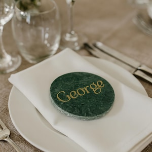 May include: A circular, green marble place card inscribed with the name "GEORGE" in gold lettering. The place card sits on a white napkin on a white plate, with cutlery and glasses visible. Suitable for weddings or formal occasions.
