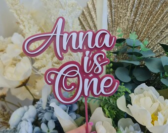Puede incluir: Un adorno para pastel rosa y blanco con el texto "Anna is One" en una fuente cursiva. El adorno está hecho de acrílico y es perfecto para una fiesta de primer cumpleaños.