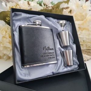 May include: A black leather-covered hip flask with a silver trim and engraved text: "Nathan,  Groomman, Mr & Mrs Smith, 19.06.2020". The flask is in a black box with a grey satin lining. Two silver shot glasses are included.