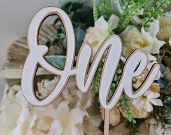 Puede incluir: Un adorno para tarta de madera con la palabra "One" en cursiva. La palabra es blanca con un borde marrón claro y un palito de madera. El fondo presenta arreglos florales blancos y verdes. Perfecto para un primer cumpleaños.