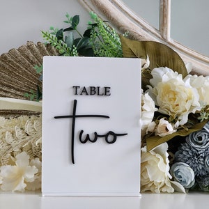 May include: White acrylic table number sign with black text that reads "TABLE TWO" in a cursive font.