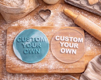 May include: A wooden cutting board with two round objects that read "CUSTOM YOUR OWN". One is blue, the other is clear. Baking supplies like flour, a rolling pin, and a whisk are scattered around the board.