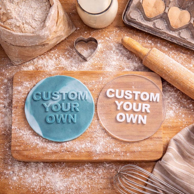 Puede incluir: Una tabla de cortar de madera con dos objetos redondos que dicen "CUSTOM YOUR OWN". Uno es azul, el otro es transparente. Hay suministros para hornear como harina, un rodillo y un batidor.