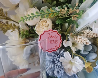 May include: A clear glass with a floral arrangement in the background. The flowers are white, cream, and blue. A pink tag with the text "Tara's HENS ANNALISE" is in front of the glass. The arrangement includes greenery and various flower types.
