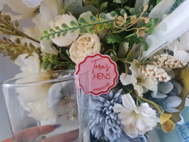 Puede incluir: Un vaso transparente con un arreglo floral en el fondo. Las flores son blancas, crema y azules. Una etiqueta rosa con el texto "Tara's HENS ANNALISE" est&aacute; delante del vaso. El arreglo incluye vegetaci&oacute;n y varios tipos de flores.