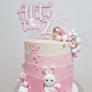 Pode incluir: Um bolo de dois andares com um design gradiente rosa e branco. O bolo é decorado com uma figura de coelho, balões rosa e dourados e um topo de bolo que diz "Alita twins 2".