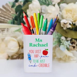 May include: White ceramic pencil holder with the text "Ms Rachael You just hap-pen to be just ink-credible" and a red apple illustration. The holder is filled with colorful pens and pencils.