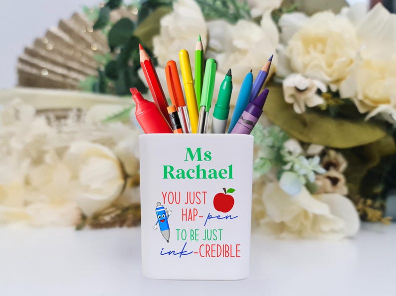 May include: White ceramic pencil holder with the text "Ms Rachael You just hap-pen to be just ink-credible" and a red apple illustration. The holder is filled with colorful pens and pencils.