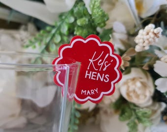 May include: A red, scalloped-edge drink charm with white text that reads "Kels Hens" and "Mary". The charm is attached to a clear glass, with a blurred floral arrangement in the background.