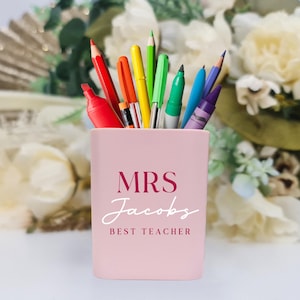 Puede incluir: Un portalápices rosa claro con el texto "MRS Jacobs BEST TEACHER" en burdeos y blanco. Contiene varios lápices de colores, bolígrafos y rotuladores. El fondo presenta flores blancas y amarillas.
