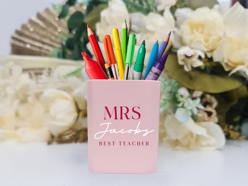 May include: Pink pencil holder with the text "MRS Jacobs BEST TEACHER" in burgundy lettering. The holder is filled with a variety of colorful pens and pencils.