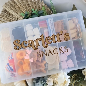 May include: A clear plastic container with compartments filled with various snacks. The container has a label that says "Scarlett's Snacks".