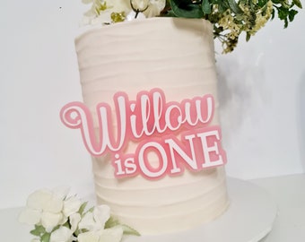 Puede incluir: Un pastel blanco con glaseado rosa y una decoración de acrílico rosa y blanco que dice "Willow is ONE".