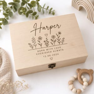 Puede incluir: Una caja de recuerdos rectangular de madera clara con un cierre de metal. La tapa presenta el nombre Harper en cursiva, una línea de ilustraciones florales negras y el texto BORN WITH LOVE CHERISHED ALWAYS seguido de la fecha 12.06.2024.