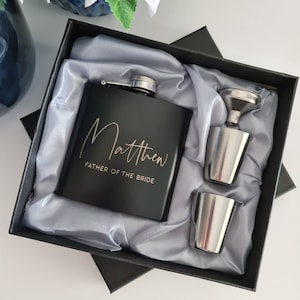 May include: A black hip flask gift set, including a silver funnel and two shot glasses, presented in a black box with grey satin lining. The flask is engraved with the name "Matthew" and "Father of the Bride".