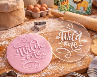 Może przedstawiać: Różowy, okrągły fondant z białym napisem "Wild one" i wzorem strzałki. Widoczny jest również przezroczysty, okrągły topper na ciasto z tym samym tekstem. Składniki do pieczenia, w tym jajka i mąka, znajdują się w tle.