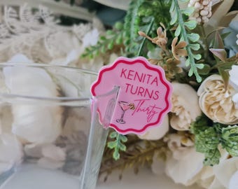 May include: A pink drink charm with scalloped edges, featuring the text "KENITA TURNS Thirty" and a cocktail illustration. The charm is attached to a clear glass, with floral arrangements in the background.