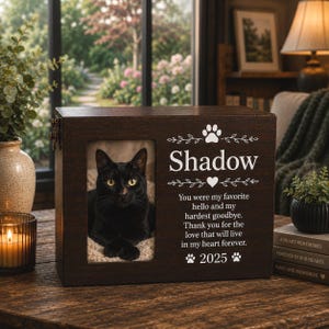 May include: A dark brown wooden pet memorial box featuring a rectangular photo frame containing a black cat. The box is engraved with the name Shadow, a paw print, a heart, and the text: You were my favourite hello and my hardest goodbye. Thank you for the love that will live in my heart forever. 2025.