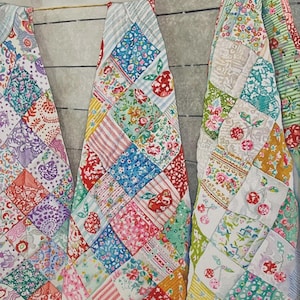 May include: Three colorful patchwork quilts hanging. Each quilt features a variety of floral and patterned fabrics in shades of red, blue, green, and yellow. The quilts are made of small, square fabric pieces sewn together in a diamond pattern.