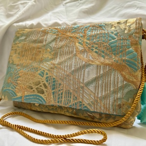 Può includere: Una pochette blu e oro con una tracolla a catena di corda dorata. La borsa ha una chiusura a lembo e un pon pon.