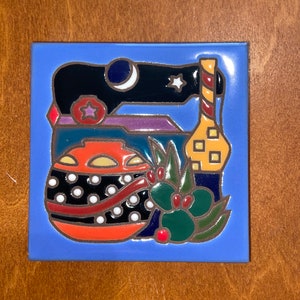 May include: A square ceramic tile with a blue background features a colorful illustration. The design includes a black shape with a crescent moon and star, a bottle, a round ornament with white dots, and holly with red berries.