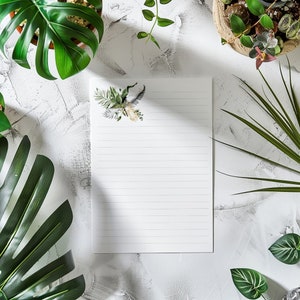 May include: A white lined notepad with a floral design on the top left corner. The notepad is on a white surface with green plants around it.