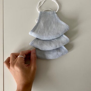Puede incluir: Tres mascarillas de lino azul claro con elásticos de oreja blancos. Las mascarillas están apiladas una encima de la otra.