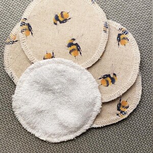May include: A set of reusable makeup remover pads. The pads are made of soft, white fabric and are stacked on top of each other. The top pad is a white cotton pad, and the bottom pads are made of a beige fabric with a bee pattern.