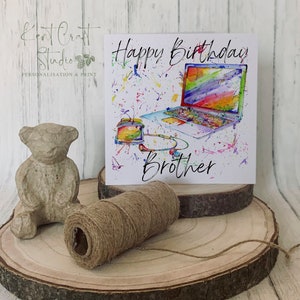 May include: A birthday card featuring a watercolour illustration of a laptop, headphones, and a paint splattered background. The card reads "Happy Birthday Brother".