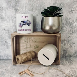 May include: White ceramic money box with a slot on the top. The money box is sitting on a wooden crate with a sign that says "Kent Craft Studio Personalisation & Print". There is a white ceramic mug with the text "Ella's Gaming Fund" and a watercolor illustration of a video game controller and flowers.