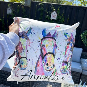 May include: White pillow with a watercolor painting of three horses' heads. The horses are painted in shades of pink, blue, green, and yellow. The pillow has the name "Annalese" printed on the bottom.