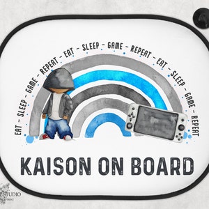 May include: A white car sunshade with a rainbow design and the text "EAT SLEEP GAME REPEAT - EAT SLEEP - GAME REPEAT" in a circle around the rainbow. The text "KAISON ON BOARD" is below the rainbow. A cartoon character is standing to the left of the rainbow. A video game controller is to the right of the rainbow.