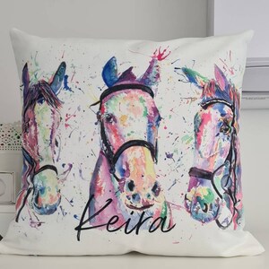May include: White pillow with a watercolor painting of three horses' heads. The horses are painted in shades of pink, blue, green, and yellow. The pillow has the name "Keira" written in black cursive.