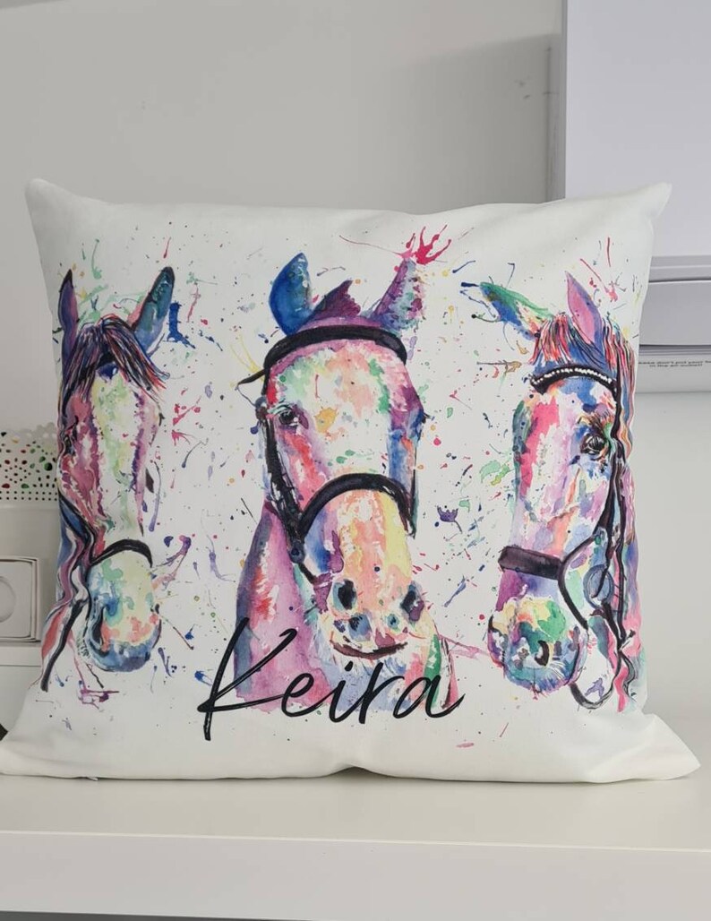 May include: White pillow with a watercolor painting of three horses' heads. The horses are painted in shades of pink, blue, green, and yellow. The pillow has the name "Keira" written in black cursive.