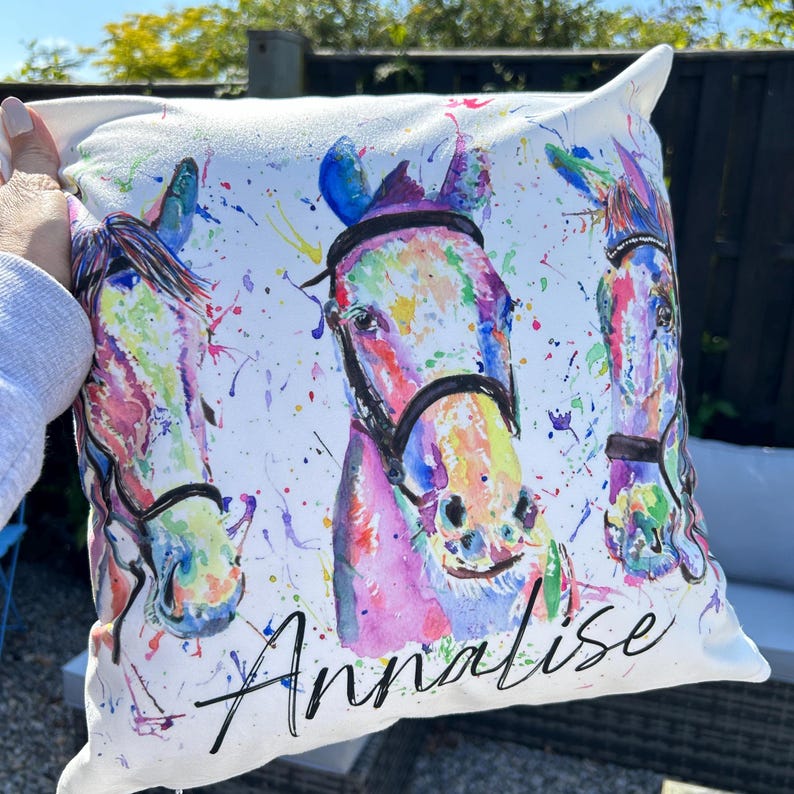 May include: White pillow with a watercolor painting of three horses' heads. The horses are painted in shades of pink, blue, green, and yellow. The pillow is personalized with the name "Annalese" in black cursive.