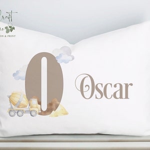 May include: A white pillow with a brown number "0" and the name "Oscar" printed on it. The number is surrounded by a watercolour illustration of a yellow cement truck, clouds, and dirt.