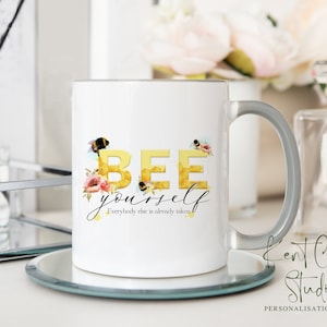 May include: White ceramic mug with a gray handle. The mug features a watercolor design of bees and flowers with the text "BEE yourself. Everybody else is already taken."