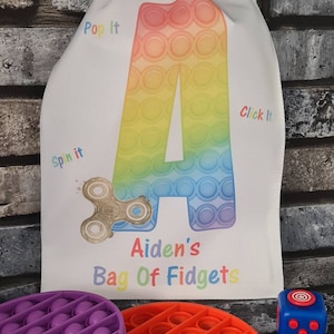 May include: A white drawstring bag with the text "Aiden's Bag Of Fidgets" and a rainbow pop it toy shaped like the letter A. The bag also has the words "Pop It", "Spin It", and "Click It" printed on it. There are three fidget toys in the image: a purple pop it, an orange pop it, and a blue fidget cube.