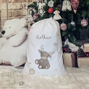 May include: A white Christmas gift bag with a drawstring closure. The bag features a cartoon reindeer illustration and the name "Arthur" in a cursive font. Gold snowflakes are scattered around the reindeer. The bag is in front of a Christmas tree.